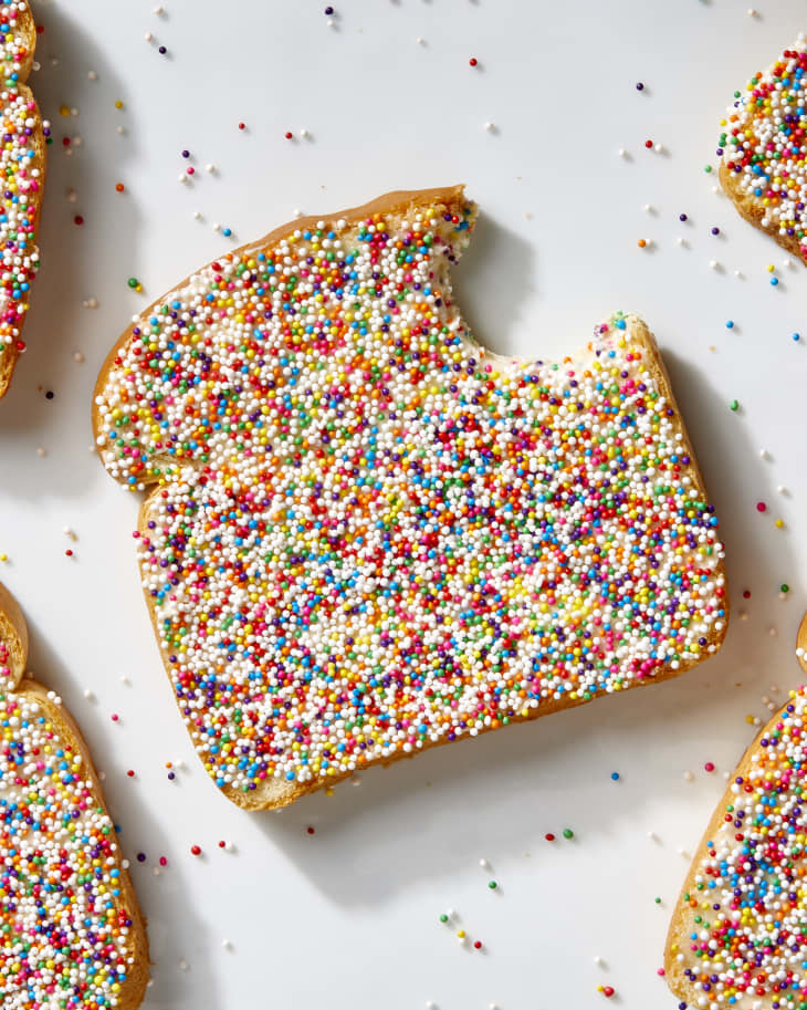 Overhead shot of a piece of fairy bread with a bite taken from the top right, on a white surface, and extra rainbow sprinkles scattered on the surface.