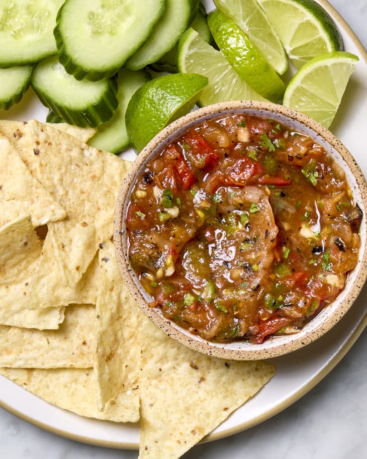 A bowl of ranchero sauce served with tortilla chips, sliced cucumber, and limes.