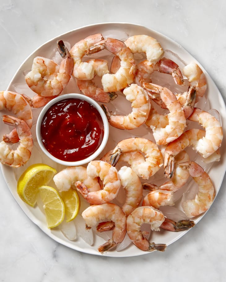 Overhead shot of cooked shrimp on ice on a large white round platter, with a small bowl of cocktail sauce in the center and some lemon wedges on the left side of the platter.