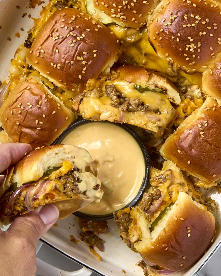 close up shot of a tray of cheeseburger sliders in a white pan, with one slider being dipped into the side of sauce on the pan.