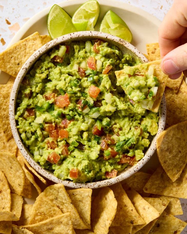 overhead shot of guacamole in a small bow surrounded by tortilla chips, and a hand holding a chip with guac on it.