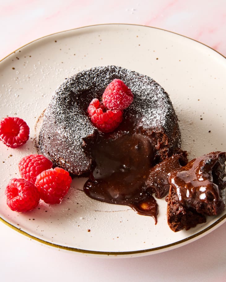 angled shot of a chocolate lava cake topped with powdered sugar and raspberries on a beige dessert plate, with a piece taken out of it, showing the gooey inside.