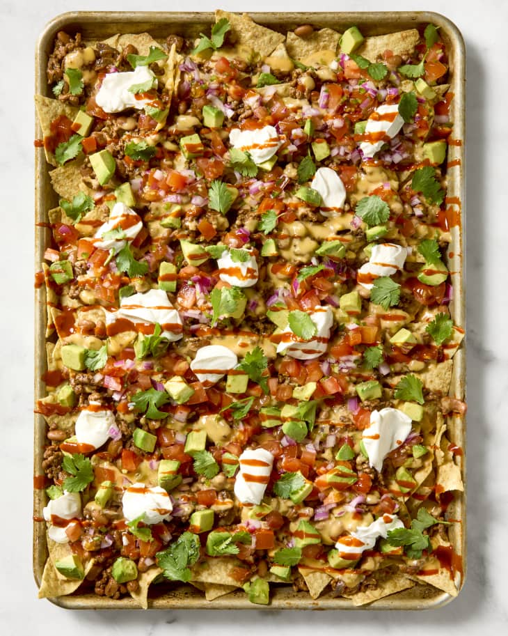 overhead shot of loaded nachos on a baking sheet, topped with sour cream dollops, avocado and a drizzle of hot sauce.