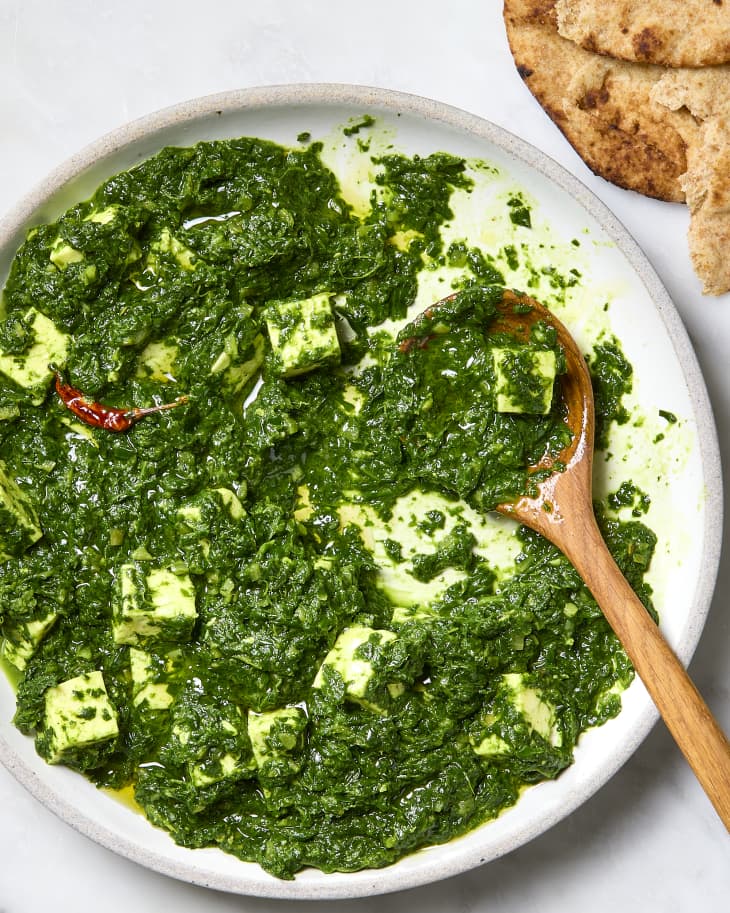 A platter of saag paneer with some food missing with a wooden serving spoon. There is whole wheat pita bread on the side.