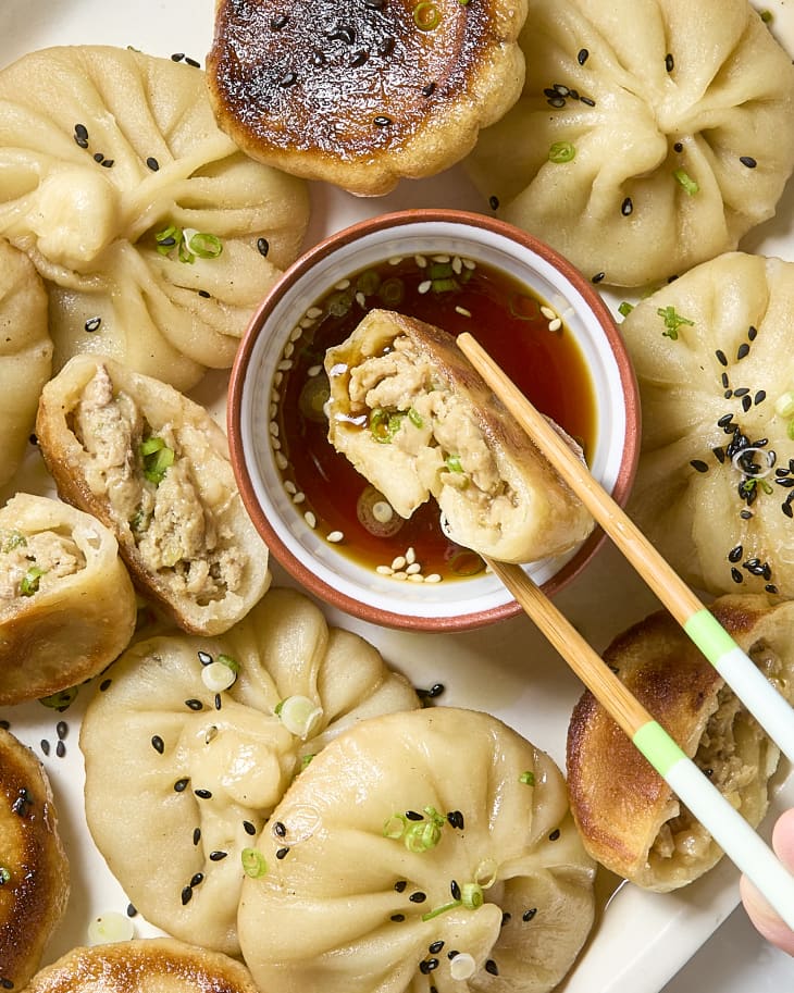 A platter of sheng jian bao (pan fried pork buns) with a small bowl containing a dipping sauce. Some of the dumplings are split in half to reveal the pork filling and one half of one is being held with chopsticks and being dipped into the sauce.