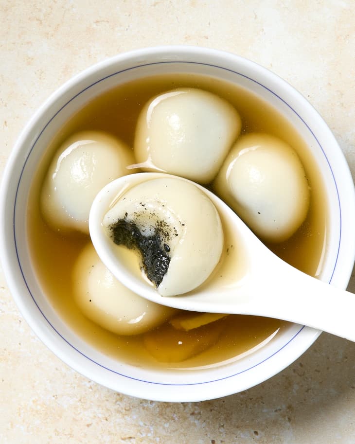 Bowl of tang yuan dessert rice balls stuffed with sesame paste in a sweet broth with one on a spoon with a bite taken out.