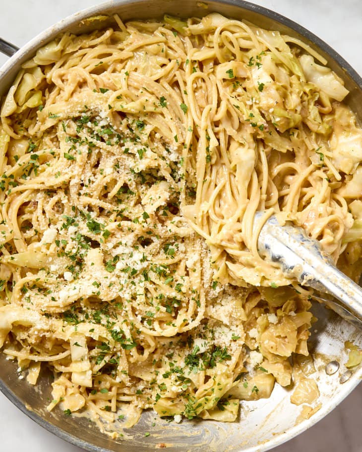 overhead shot of melty cabbage carbonara in a pan, topped with herbs and cheese - and with metal tongs grabbing a bunch of spaghetti.