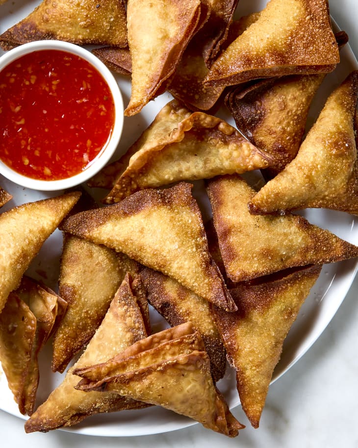 Close up shot of finished crab rangoons on a white plate with a small white bowl of sweet chili sauce to the left of them.