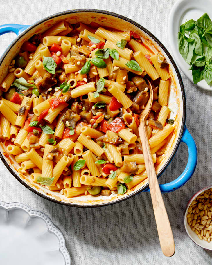 Eggplant Caponata Pasta is served in a dutch oven and sits on a textured linen next to a plate of basil and bowl of pine nuts.