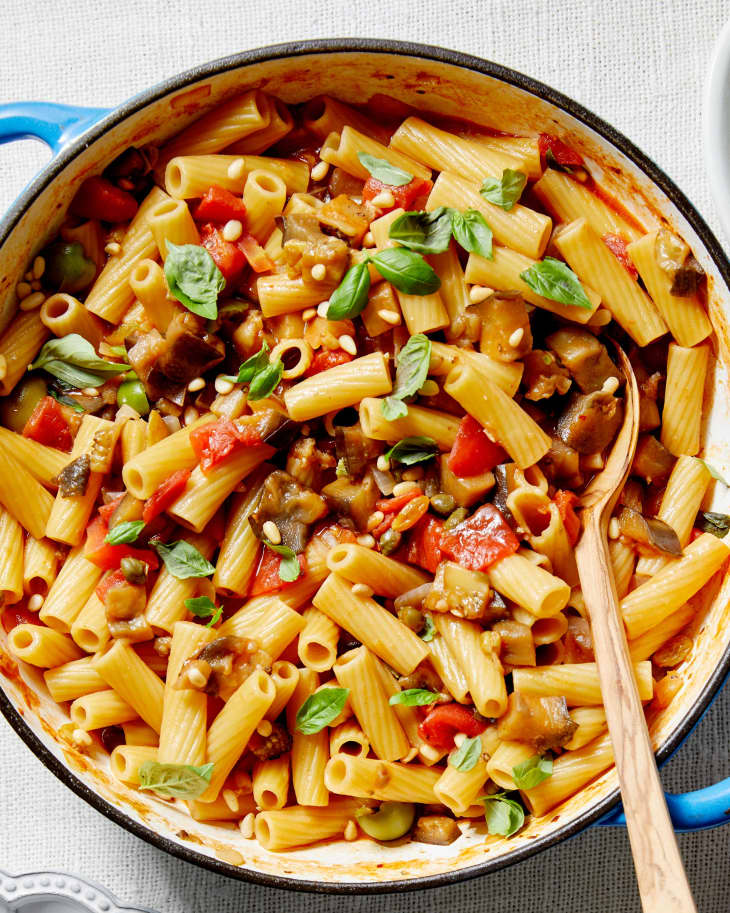 Eggplant Caponata Pasta is served in a dutch oven and sits on a textured linen next to a plate of basil and bowl of pine nuts.