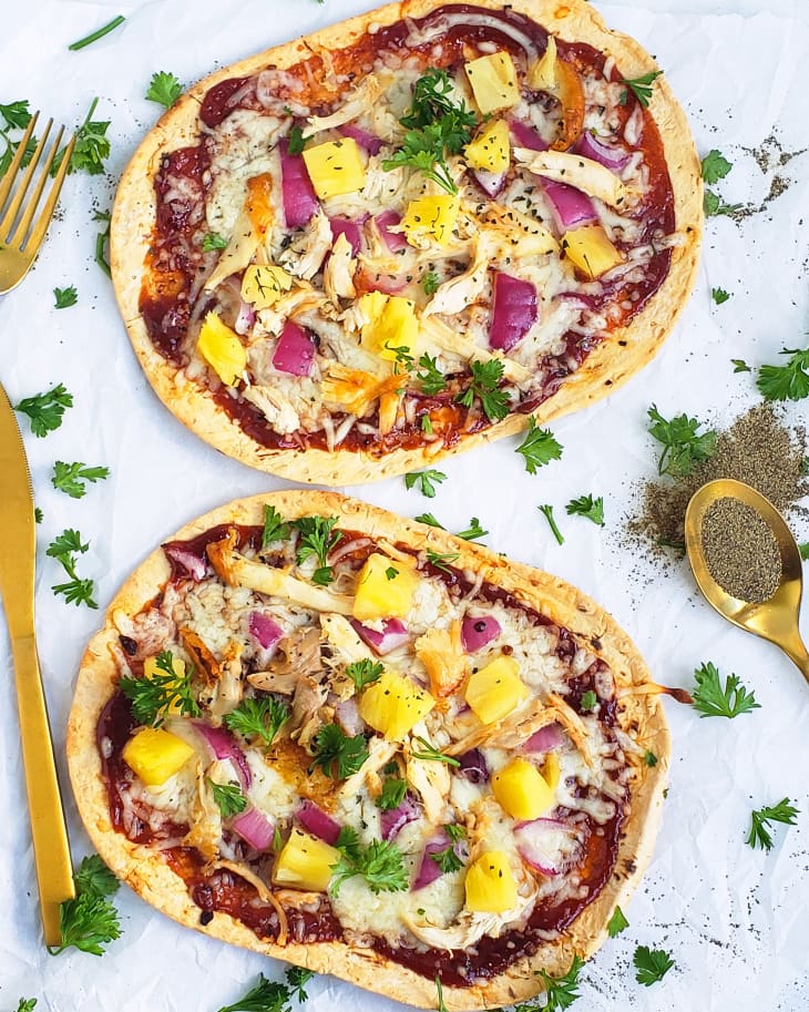 Barbecue chicken and pineapple flatbread pizza laid out on white linen with herbs sprinkled and pepper on side.