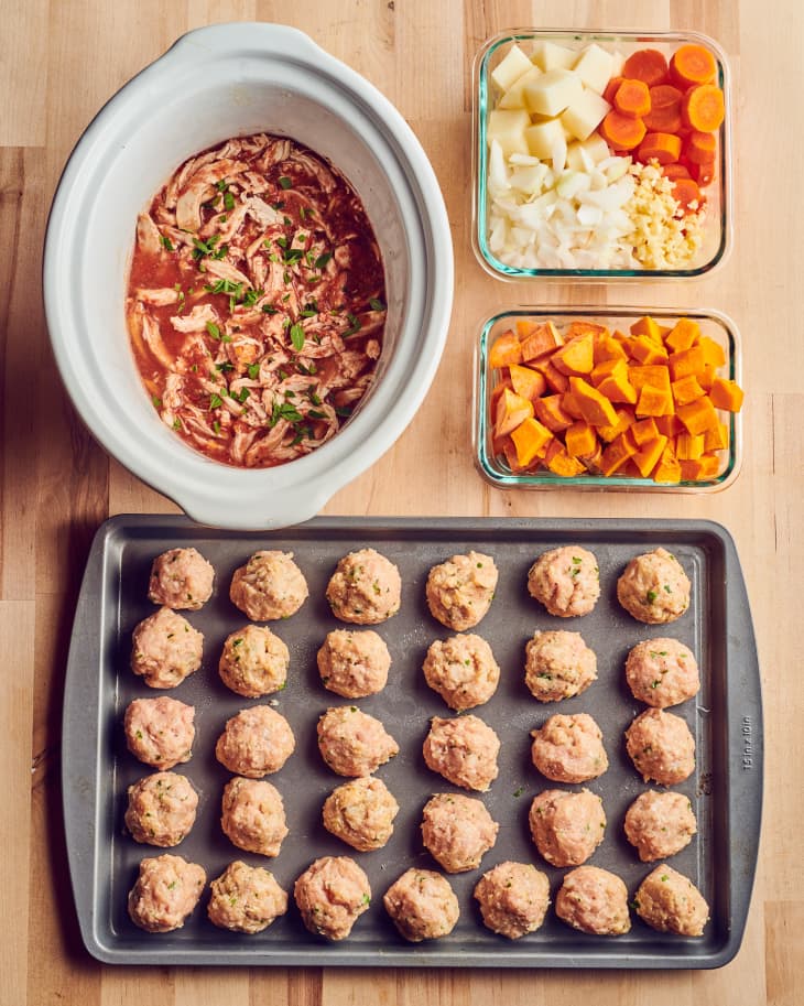 A baking sheet of formed, uncooked meatballs, a slow cooker filled with salsa pulled chicken, a par-baked squash, a sweet potato, chopped white potatoes, onion, carrots, and garlic sit on a wooden table.