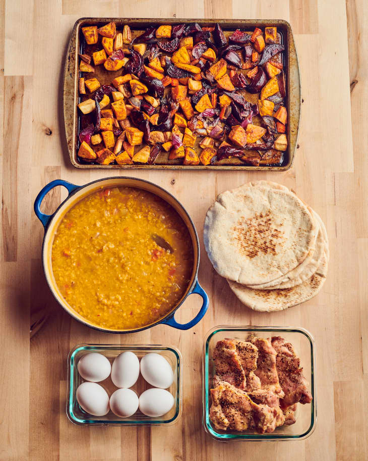 2lbs of cooked boneless chicken thighs, a sheet pan of roasted root vegetables, a bowl of hard-boiled eggs, a Dutch oven filled will lentil soup, and a stack of pita bread sit on a wooden table.
