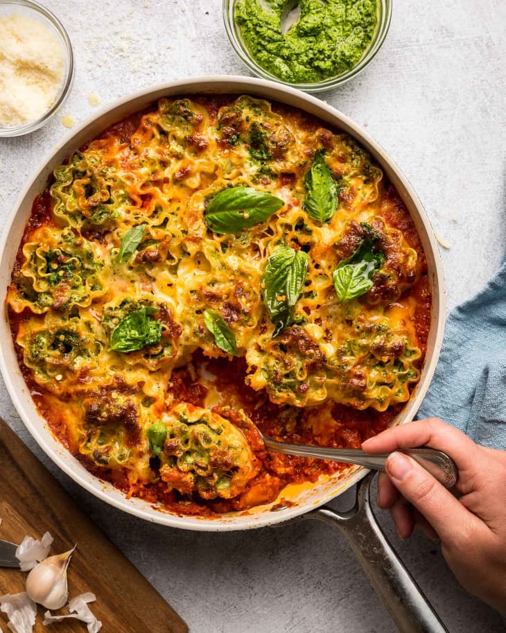 someone is stirring the skillet lasagna with a metal spoon inside of a skillet with small bowls of spices nearby and out of focus