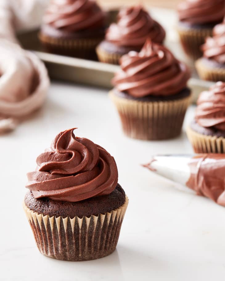 a chocolate cupcake sits on a table with a piping bag and other cupcakes behind it