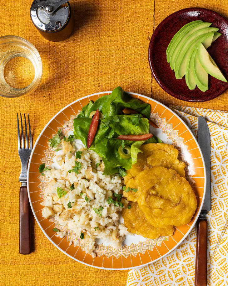 Overhead photo of plate with rice, cod, plantain tostones, and salad sitting on gold/yellow table with gold and white napkin. Bowl with sliced avocado in upper right. Pepper mill and glass of white wine