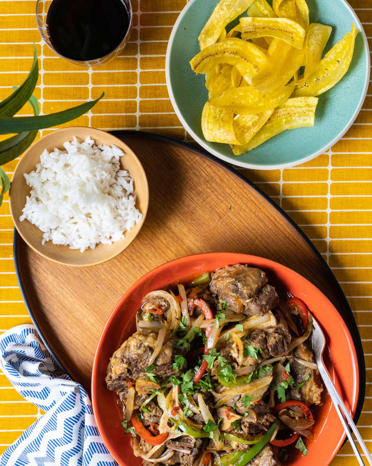 Overhead shot of stewed oxtails with peppers and onions and herbs in orange bowl. Bowl is sitting on a wood platter with a small bowl of rice next to it. There is a gold and white grid-pattern tablecloth underneath. In the upper right corner is a light aqua bowl of tostones, and in the upper left there is a glass of red wine