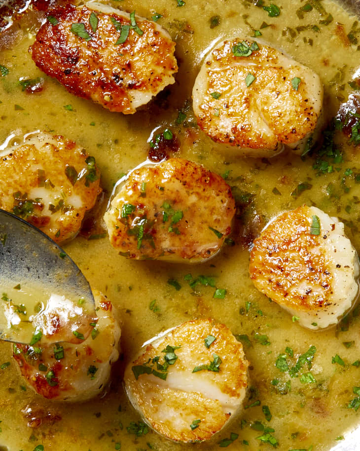 Overhead shot of 7 scallops on a sauce pan, sitting in a lemon, butter and herb sauce.