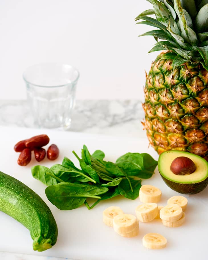 Pineapple, avocado, banana slices, spinach, zucchini, and dates on a marble surface with a glass.