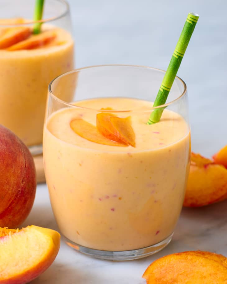 A glass of peach lassi with a straw, surrounded by cut peaches