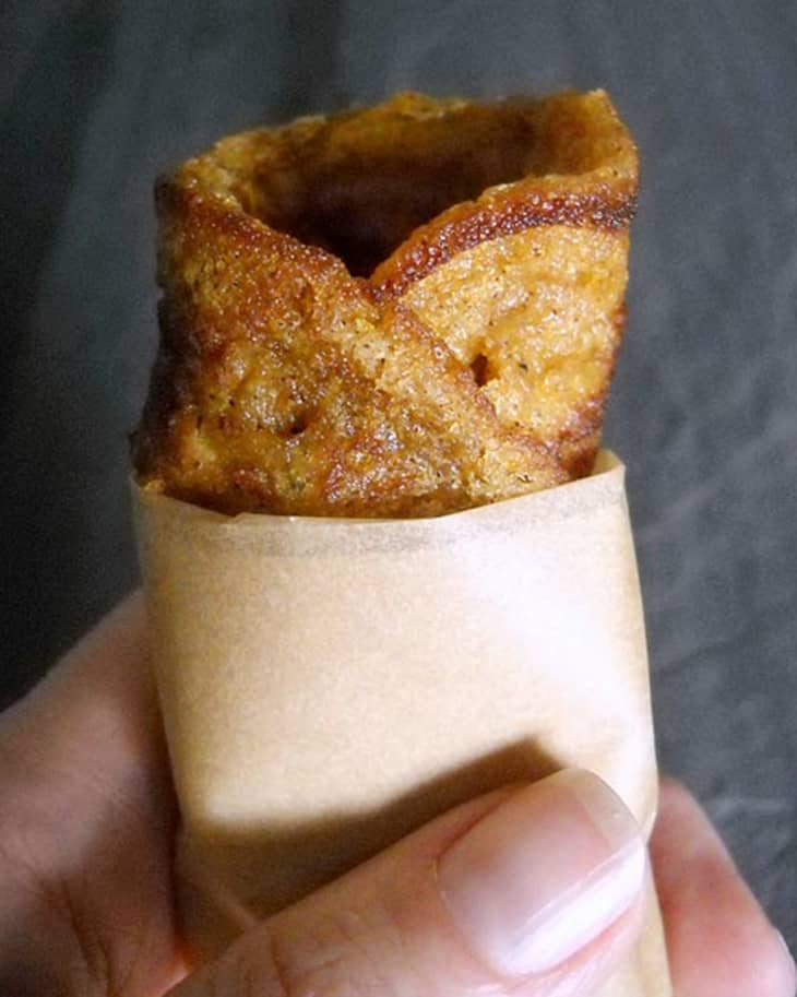 Hand holding a rolled, crispy brown snack wrapped in brown paper.