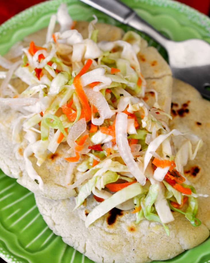 A round green plate contains masa cakes topped with freshly chopped cabbage slaw