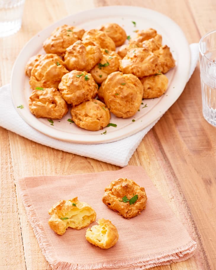 Freshly baked cheese gougères are piled up on a white plate, while a piece on a peach-colored napkin is torn to pieces