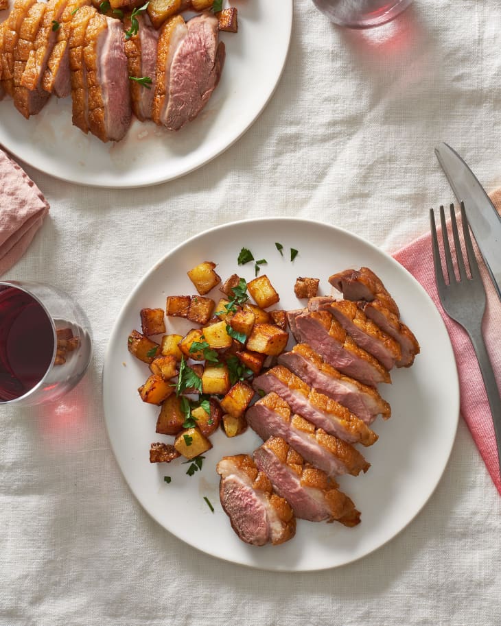 Duck breast with golden crisp skin, served with roasted potatoes and garnished with chopped cilantro, on a plate with fork and knife on the side