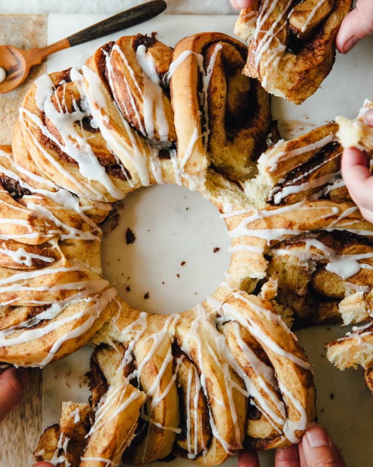 Cinnamon roll wreath with icing, being pulled apart by several hands on a white surface.