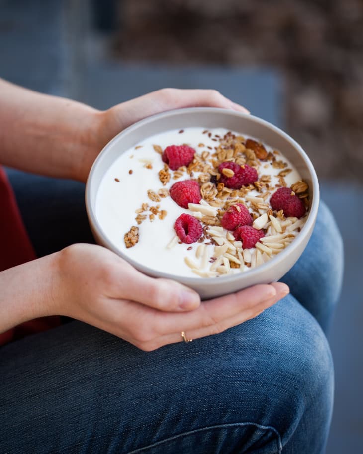 Hands holding a bowl of yogurt topped with raspberries, nuts, and seeds on a lap