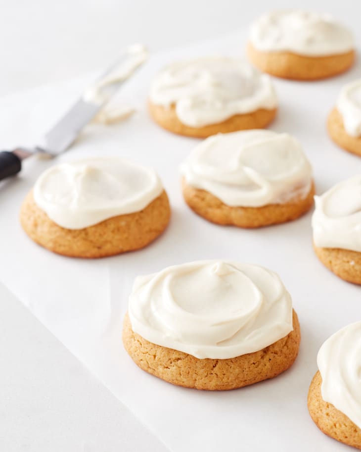 Image 3: Pumpkin cookies with creamy white frosting on parchment paper, next to a frosting spatula.