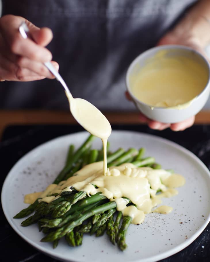 Spooning Hollandaise sauce from a small bowl over a plate of cooked asparagus