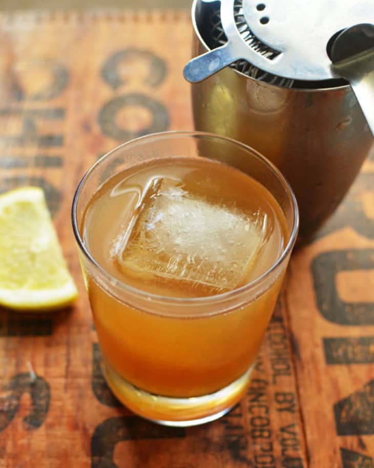 A cocktail with ingredients of honey, lemon and bourbon, with an ice cube