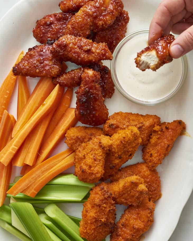 Someone dips the baked boneless chicken wings in a dressing on a plate, with slices of carrots and celery on the side