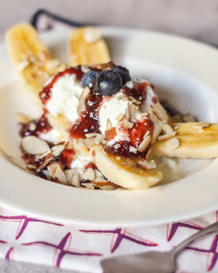 Banana split with yogurt, jam, sliced almonds, and blueberries on a white plate.
