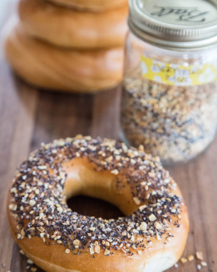 A bagel topped with everything bagel seasoning mix is pictured against a stack of plain bagels and a mason jar filled with seasoning mix