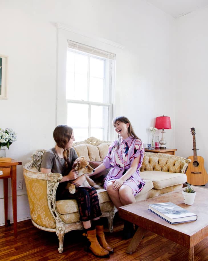 Two women sitting on a vintage sofa with a dog, next to a wooden coffee table and a guitar in a bright living room.