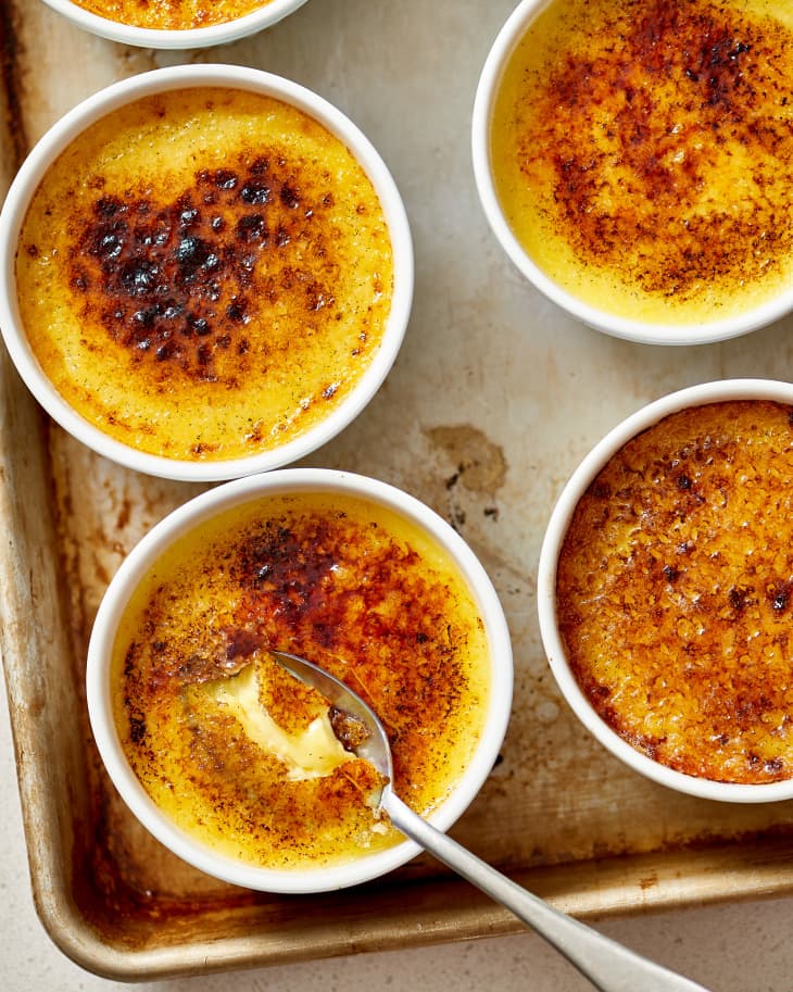 Several ramekins of creme brulee on a baking sheet, a spoon diving into one
