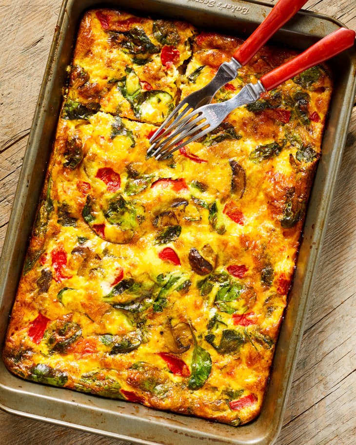 A breakfast casserole, baked with spinach, peppers, potatoes, and mushrooms, in a baking pan with two forks on it