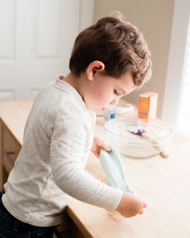 A young child in a white long-sleeve shirt is holding a blue towel while standing at a wooden kitchen counter with a glass bowl and ingredients nearby.