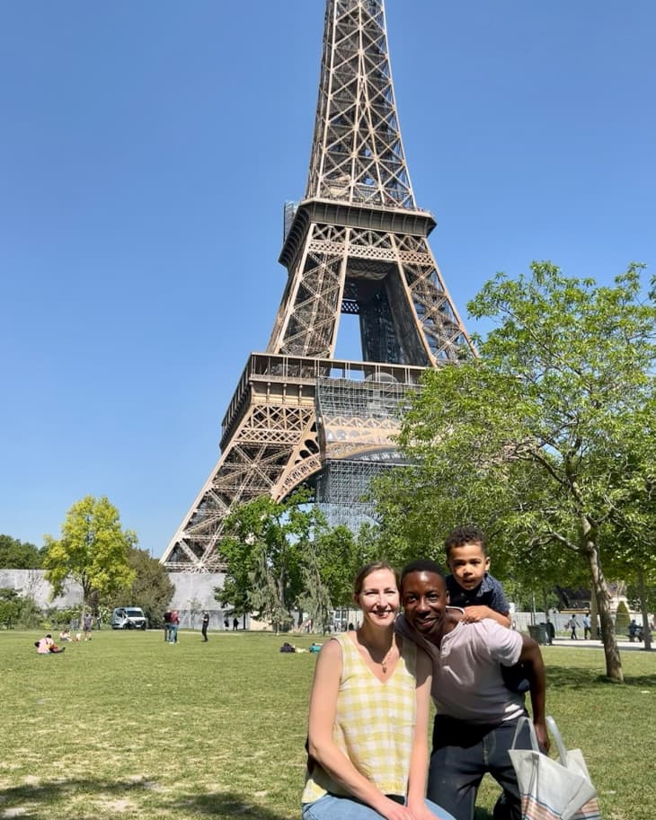 Family sitting on grass in front of the Eiffel Tower, with trees and people in the background.