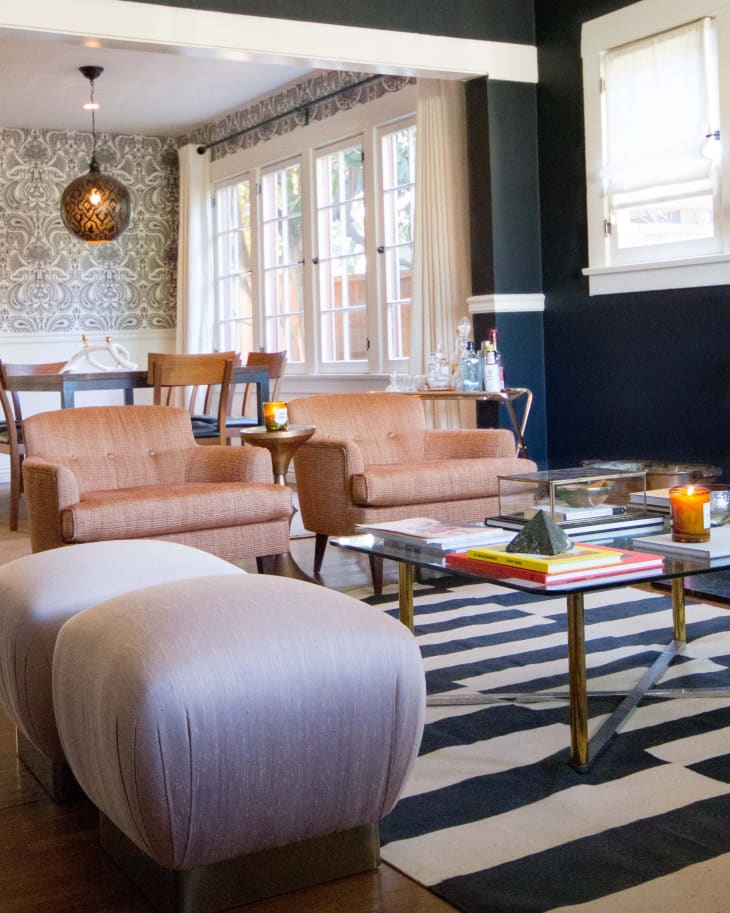 Living room with patterned wallpaper, beige armchairs, striped rug, glass coffee table, and a dining area in the background.