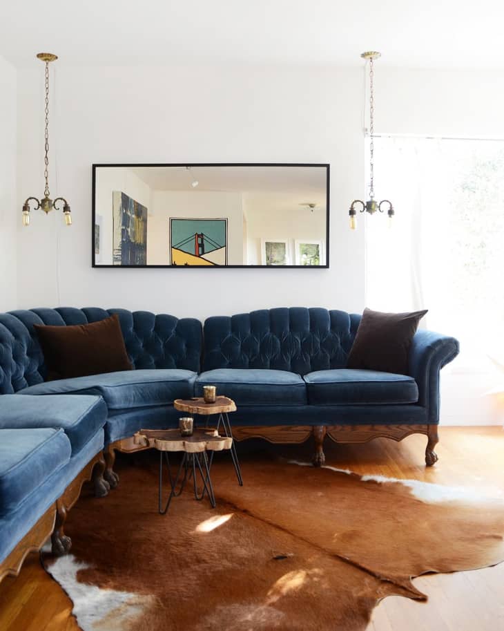 Blue velvet tufted sectional sofa with brown pillows, cowhide rug, wooden side tables, and pendant lights in a living room.