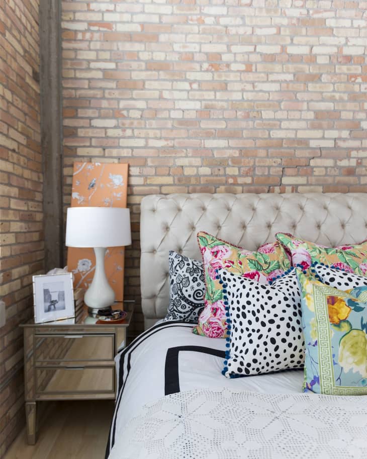 Tufted headboard with colorful patterned pillows, mirrored nightstand, white lamp, and exposed brick wall.