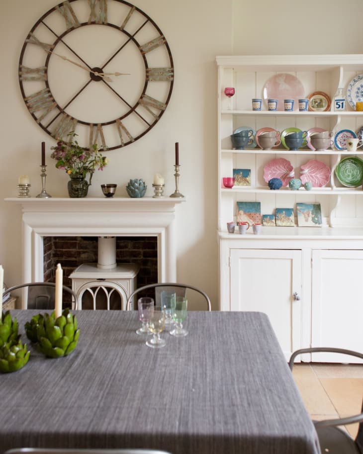 Dining room with large wall clock, gray tablecloth, candles, and a white cabinet displaying colorful dishes.