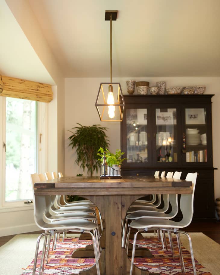 Dining room with a wooden table, white chairs, pendant light, colorful rug, and a dark cabinet with decorative items.