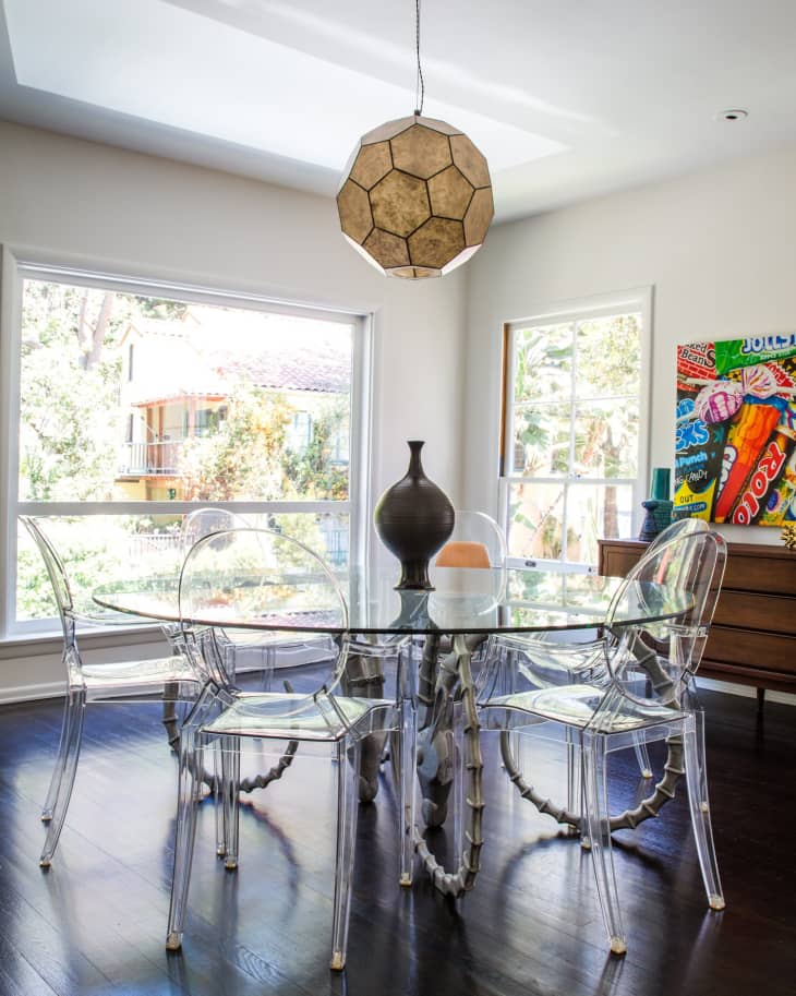 Glass dining table with clear chairs, geometric pendant light, and colorful wall art in a bright room.