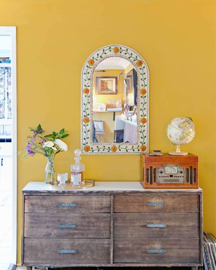 Wooden dresser with vintage radio, globe, and floral mirror on a yellow wall. Vase with flowers and glass decanter on top.