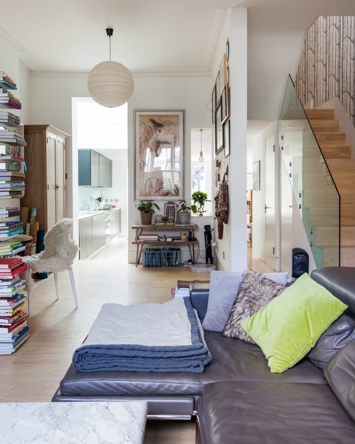Modern living room with gray leather sofa, colorful cushions, bookshelves, and view into a bright kitchen.