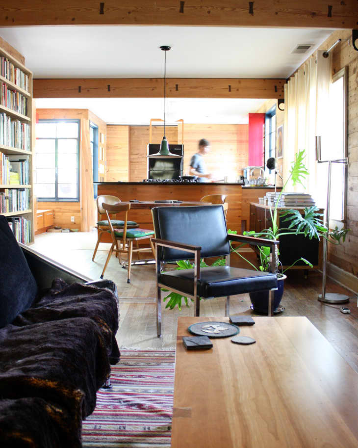 Open-plan living room with black leather chairs, wooden table, bookshelves, and a person in the kitchen area.